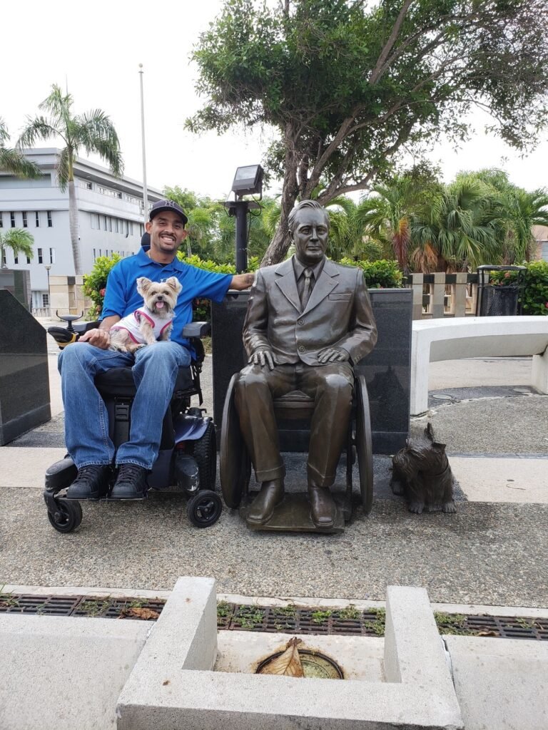 A man in a wheelchair wearing a blue polo shirt and baseball cap sits next to a bronze statue of a man in a business suit. The man holds a small white dog with brown markings. The setting appears to be outdoors with palm trees and a white building visible in the background, suggesting a warm climate location. Another small dog is visible on the ground near the statue.