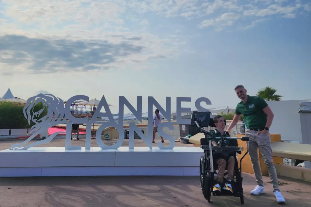 Two people at what appears to be the Cannes Lions festival or conference, as indicated by the large "CANNES LIONS" sign behind them. One person is seated in a power wheelchair, and another person in a green shirt stands behind them. The setting appears to be an outdoor terrace or venue area with palm trees and festival infrastructure visible.
