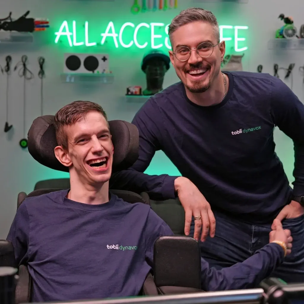 Two people in what appears to be a modern accessibility technology center or clinic. One person is seated in a power wheelchair wearing a dark blue shirt, while another person wearing glasses and a dark shirt with "tobii dynavox" branding stands behind them. There's a green neon "ALL ACCESS" sign visible in the background along with various accessibility equipment.