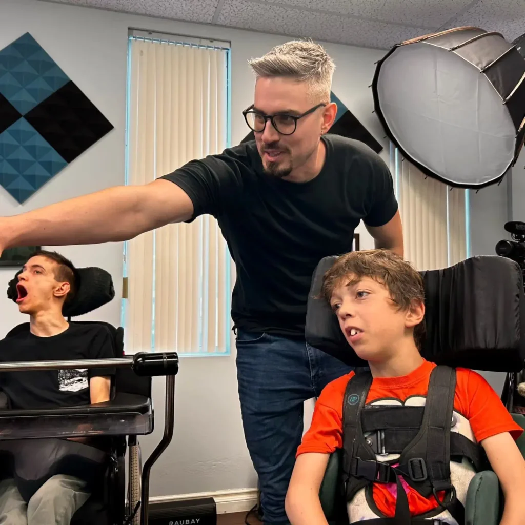 Three people in what appears to be a therapy or clinical setting with professional lighting equipment. One person is seated in a power wheelchair on the left wearing a checkered shirt, a man with glasses and dark shirt stands in the center, and another person is seated in a wheelchair on the right wearing an orange shirt. There are geometric blue and black wall decorations visible in the background.