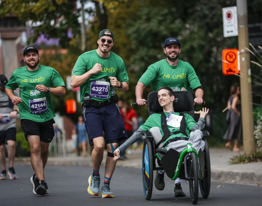 Four people participating in what appears to be a marathon or running event on a street. Three people are running alongside one person who is in a racing wheelchair. All are wearing matching green "NO LIMITS" t-shirts and race numbers. The person in the wheelchair is wearing race number and appears to be actively participating in the race. There are trees and urban surroundings visible in the background.