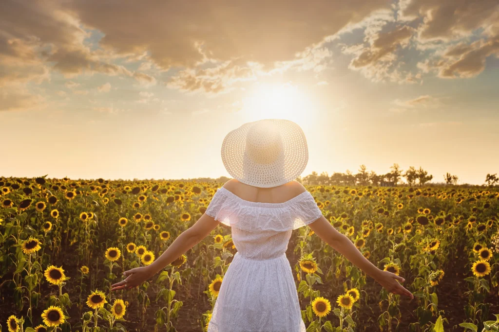 A woman in a white dress and wide-brimmed hat stands in a sunflower field, arms outstretched, as the sun sets behind her.