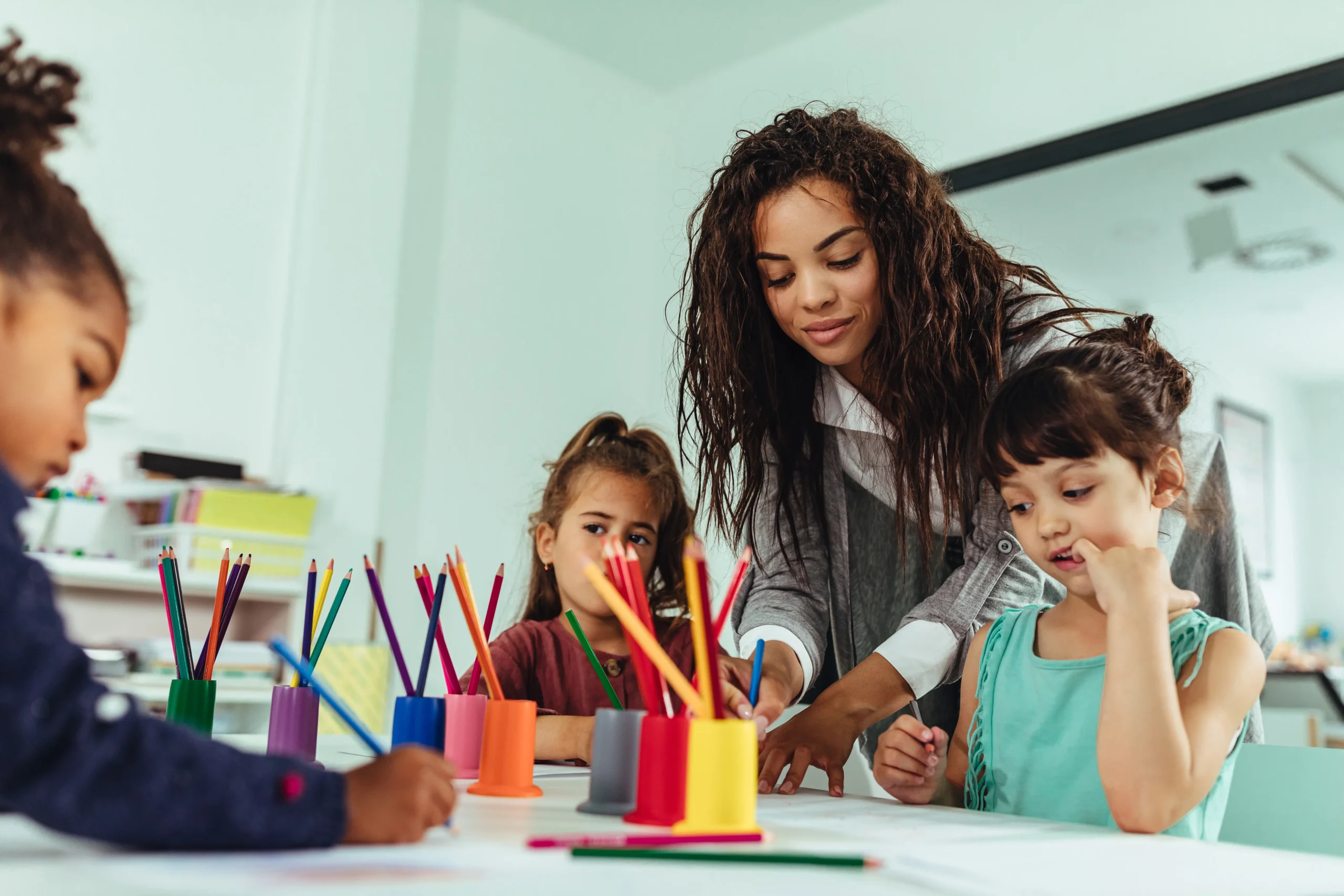 A teacher with long, curly dark hair leans over a table, helping a young girl color. Two other children are also at the table, drawing with colored pencils. Various colorful pencil holders are on the table.