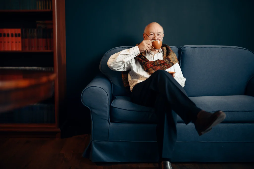 A smiling elderly man with glasses, wearing a striped shirt and a patterned scarf, sits comfortably on a blue sofa while holding an orange cup to his mouth. A wooden bookshelf filled with books is visible on the left.