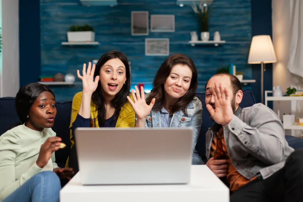 Four people, two women and two men, are gathered around a laptop on a white coffee table, waving and smiling at someone on a video call. The background shows a living room with a blue textured wall and shelves.