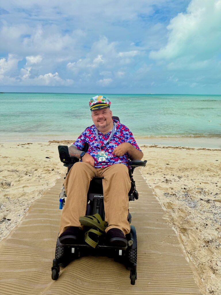 Man in a colorful shirt and cap smiles in a wheelchair on a sandy beach path, with turquoise ocean and cloudy sky in the background.