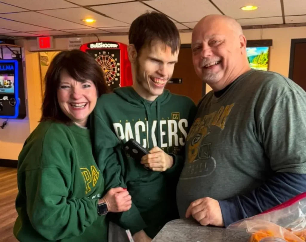 Three people posing together in what appears to be an arcade or entertainment venue. A woman on the left wearing a green Packers sweatshirt, a young man in the middle wearing a green Packers hoodie, and an older man on the right wearing a gray shirt. All three are smiling at the camera. There's a dartboard visible in the background.
