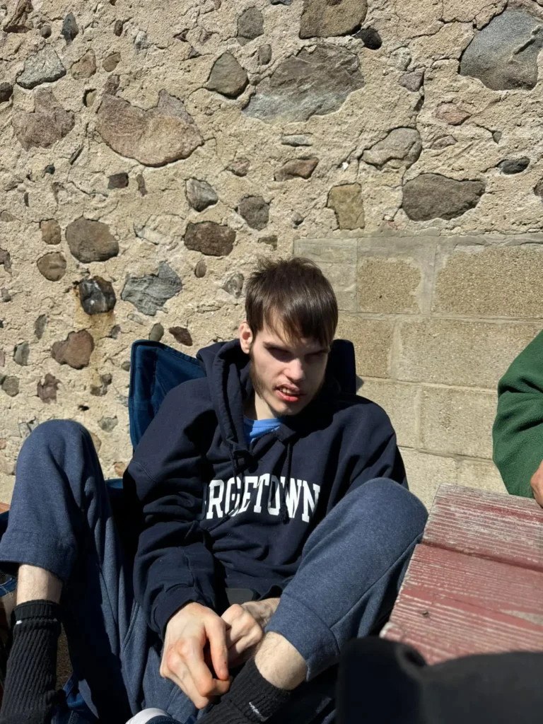 A young man with dark hair sitting outdoors against a stone wall, wearing a dark blue hoodie with "ROETOWN" text. He appears to be looking down, and there are other people partially visible in the frame.