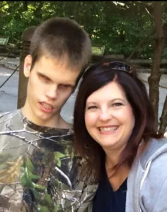 A young man and woman taking a selfie outdoors. The young man is wearing a camouflage shirt and the woman is wearing a gray hoodie. Both are smiling at the camera with trees and natural scenery in the background.