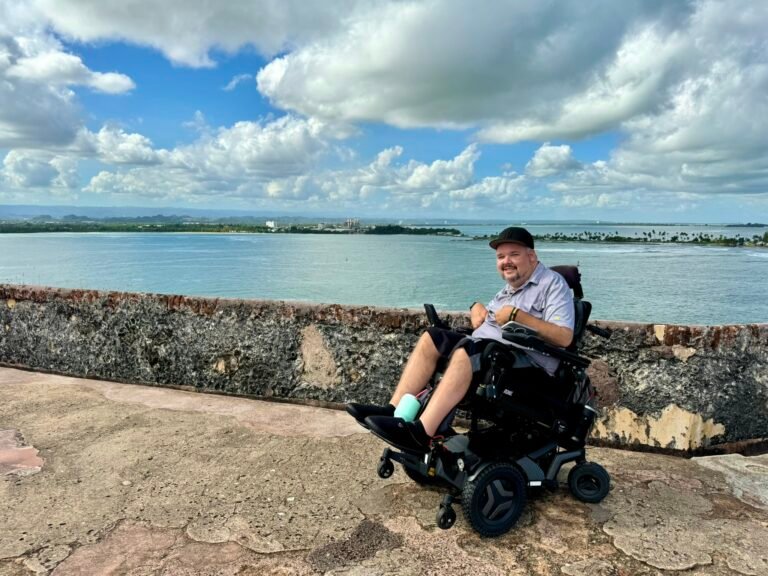 A smiling man in a power wheelchair sits on a stone walkway overlooking calm water and a distant shoreline under a blue sky with puffy clouds.