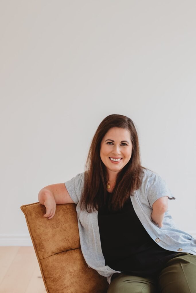 A professional headshot of a woman with long brown hair wearing a light blue cardigan over a black top, sitting on a tan leather chair against a white background