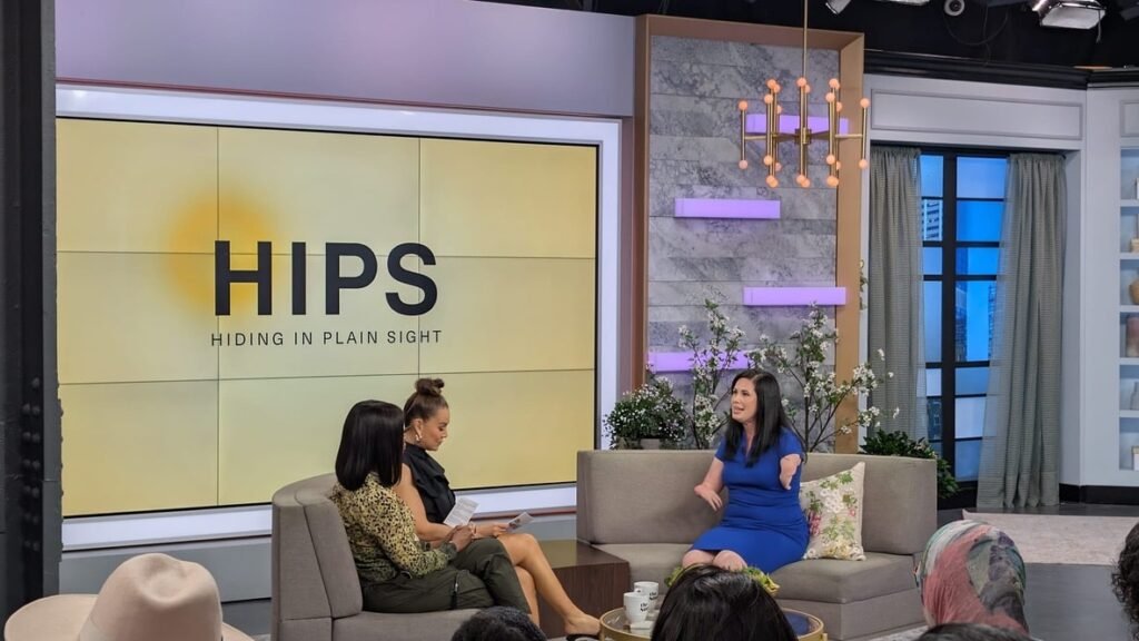 Two women sitting on a gray couch during what appears to be a TV interview, with 'HIPS - Hiding in Plain Sight' displayed on screens behind them in a modern studio setting