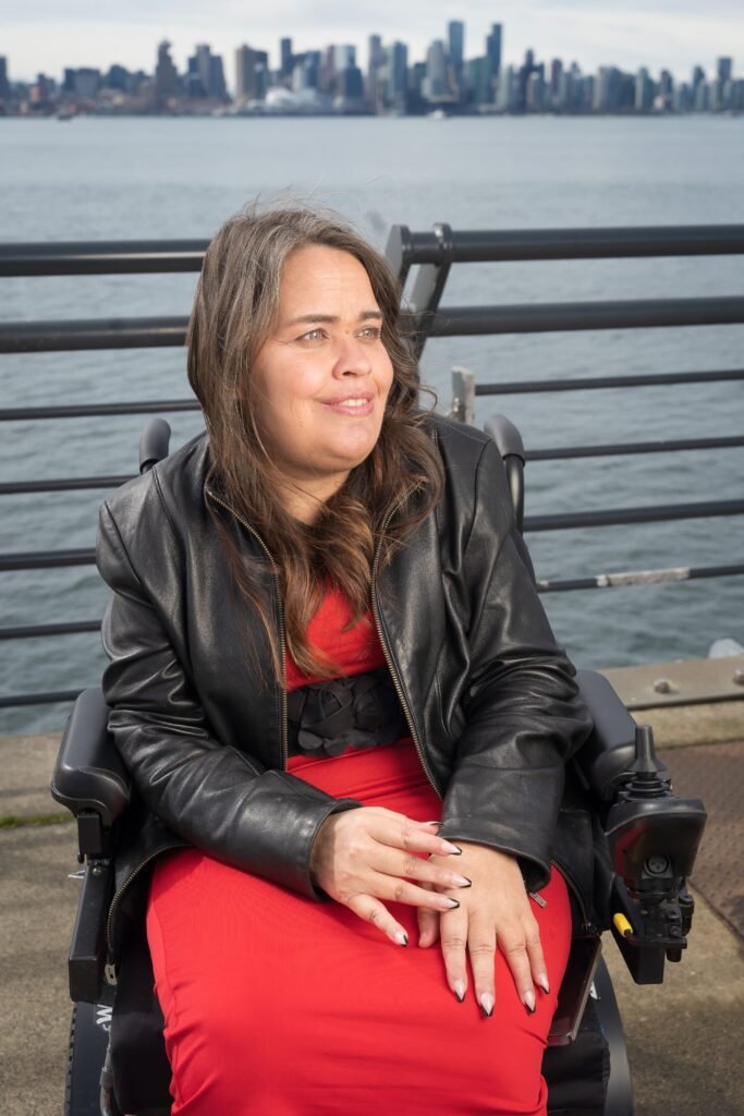 A woman in a black leather jacket and red dress sits in a wheelchair on a pier, smiling with a city skyline and water in the background.