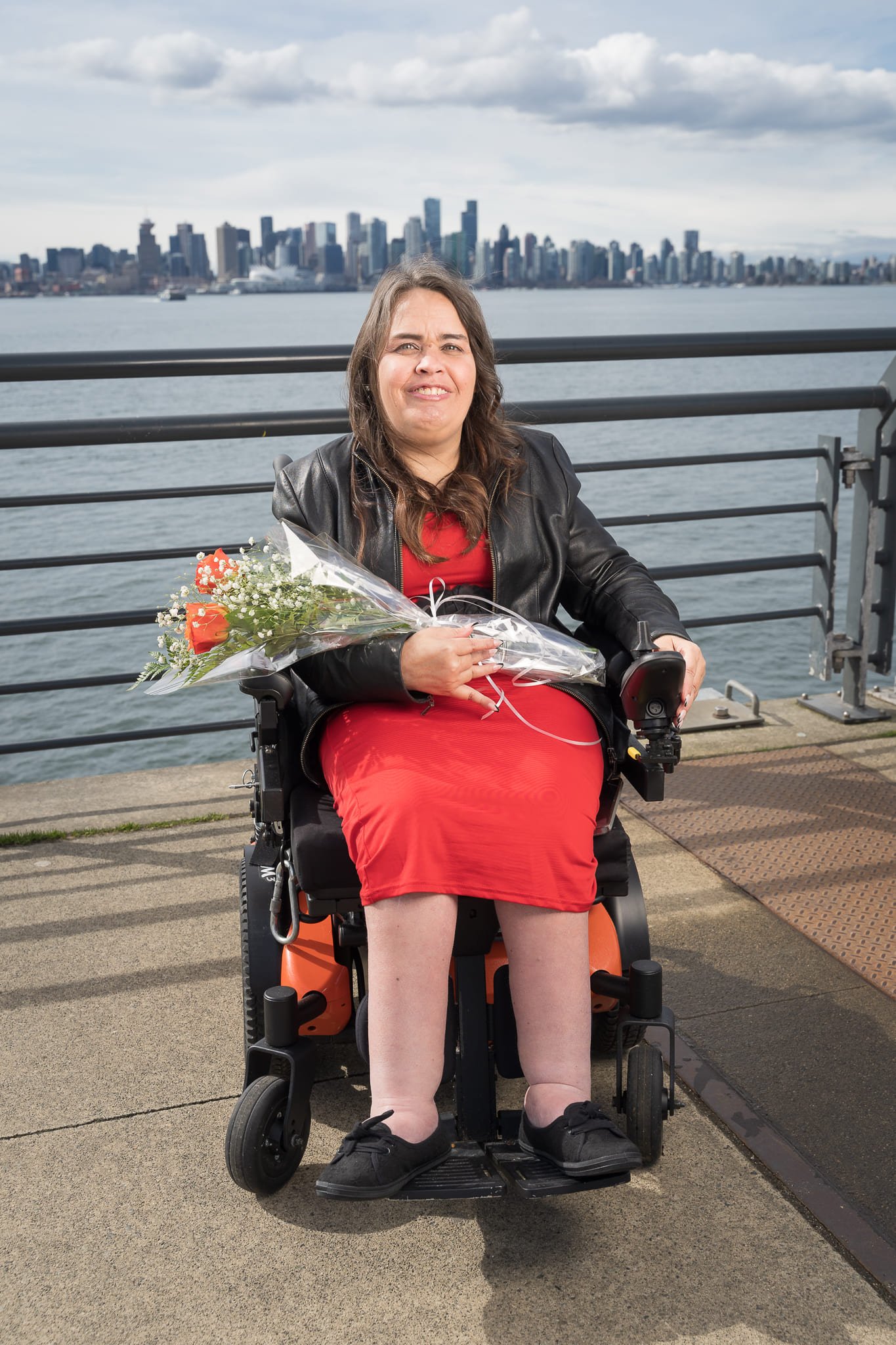 A woman in a red dress and black leather jacket sits in a wheelchair holding flowers. She smiles against a city skyline by the water on a cloudy day.