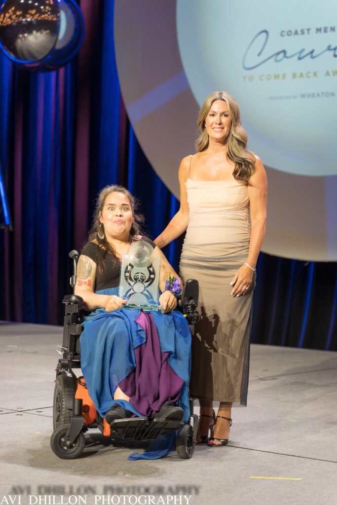 Two women on stage, one in a wheelchair holding an award, both smiling. Behind them is a large screen with text. The atmosphere is celebratory.