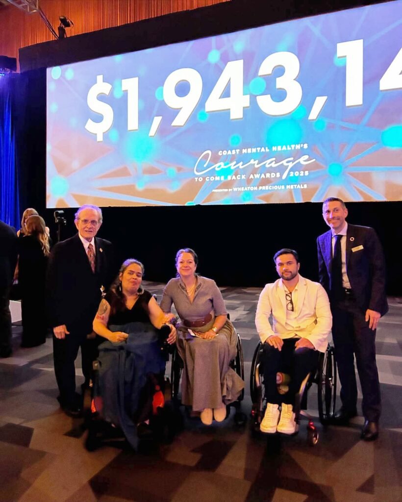 A group of people, including two in wheelchairs, smile warmly in front of a large screen displaying the number $1,943,140 at a formal event.