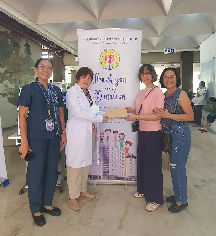 Two women in casual attire and a doctor in white coat exchange a donation in front of a banner thanking donors at the Philippine Children's Medical Center.