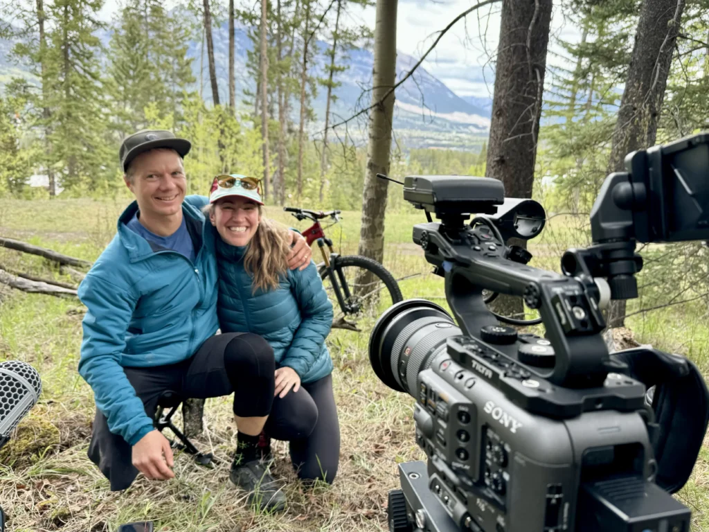 A man and woman in matching blue outdoor jackets sit close together on the ground in a wooded area, both smiling at the camera. A professional video camera is prominently positioned in the foreground, indicating this may be a behind-the-scenes moment from a film or video production. Mountain bikes are visible in the background.