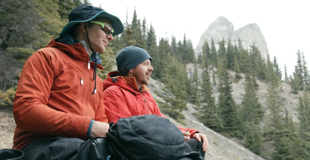 Two people in red outdoor jackets sit on rocky terrain overlooking a mountainous landscape. One person wears a green cap and the other a blue beanie. Behind them, snow-capped peaks and forested valleys stretch into the distance, creating a dramatic alpine backdrop.