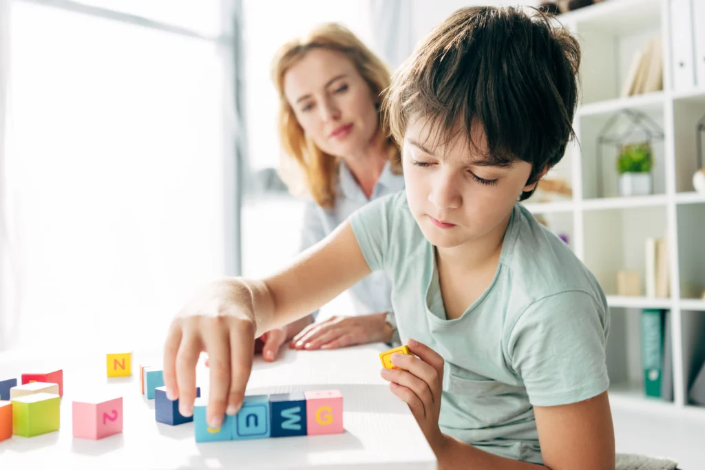 A young boy with dark hair intently focuses on stacking colorful letter blocks on a white table. An adult woman with blonde hair is blurred in the background, looking at the boy.