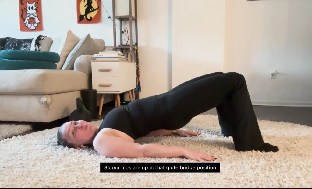 A person demonstrates a glute bridge exercise on a soft rug in a cozy living room with a couch and decorative artwork.