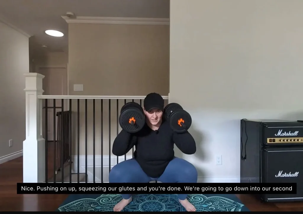 A person in workout attire performs a squat with dumbbells in a home gym, demonstrating an exercise routine on a patterned mat.
