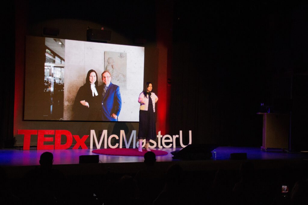 A woman giving a presentation at a TEDx McMaster University event, standing on stage next to a large screen showing a photo of two people, with the TEDx logo visible at the bottom