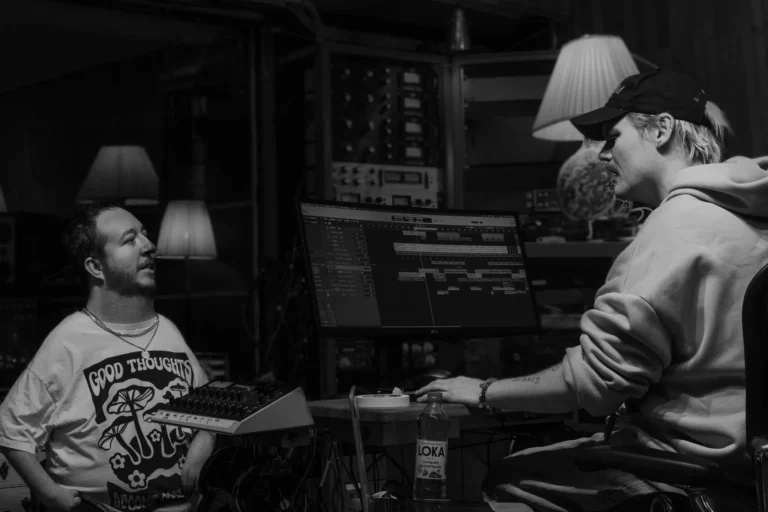Two men in a dimly lit recording studio. On the left, a man with a beard and a "GOOD THOUGHTS" t-shirt is looking at the man on the right, who is wearing a baseball cap and a hoodie, and is seated at a computer desk with a large monitor displaying music production software. A bottle of "LOKA" is on the desk.