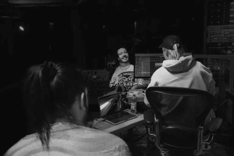 A black and white shot of three people in a recording studio. From a slightly elevated perspective, the back of a person's head and shoulders are visible in the foreground. In the midground, a bearded man in a "GOOD THOUGHTS" t-shirt is smiling, looking past the camera. To his right, another person in a cap and hoodie is seated at a computer, looking towards the bearded man. A large mixing board is visible behind them.