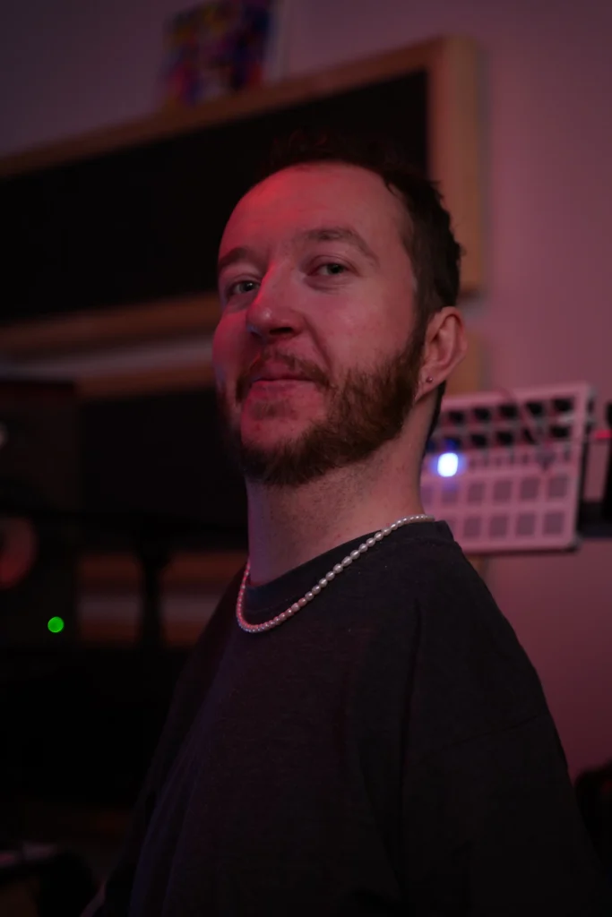 A close-up, color portrait of a bearded man with a pearl necklace, wearing a dark t-shirt. He is looking slightly to his right, with a soft smile. The background is dimly lit with a reddish hue, showing some electronic equipment.