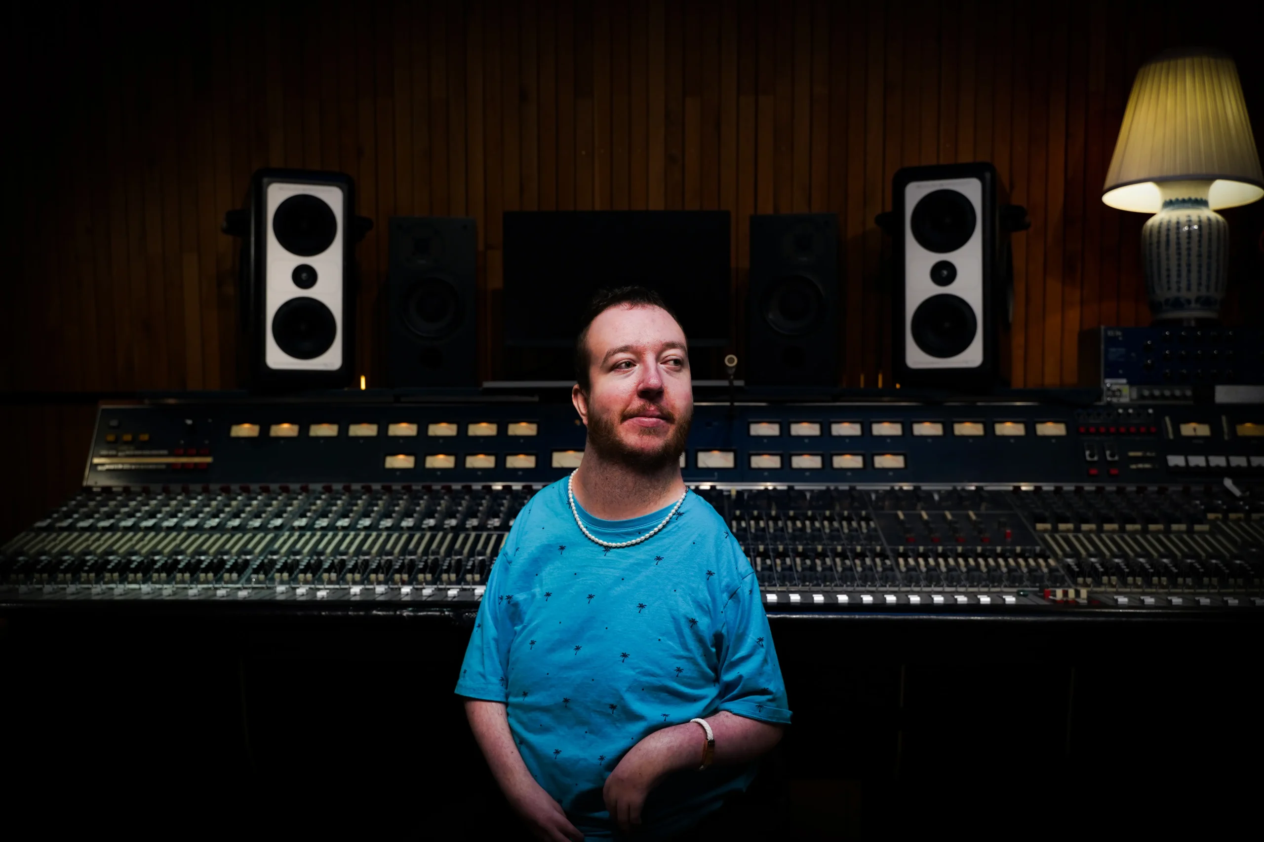 A color portrait of a bearded man with a pearl necklace, wearing a blue patterned t-shirt, seated in a recording studio. He is looking off to his right with a contemplative expression. Behind him are two large studio monitors and a mixing console. The lighting highlights him against a darker background.
