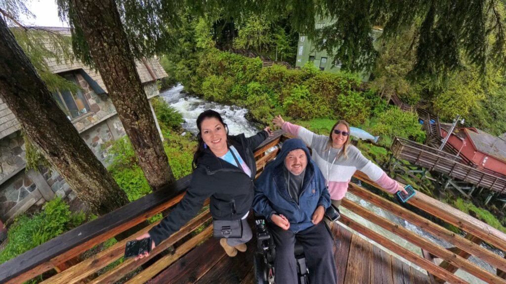Three people, one in a wheelchair, smile and spread their arms on a wooden deck in a lush, green forest area, with a stream and wooden buildings in the background.