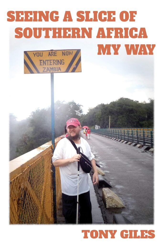 The cover of "Seeing a Slice of Southern Africa My Way" by Tony Giles. A man with a beard and a white cane smiles at the camera on a bridge. A sign on a pole behind him reads, "YOU ARE NOW ENTERING ZAMBIA."