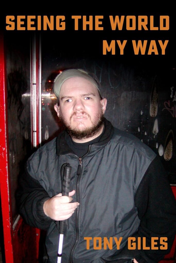 The cover of "Seeing The World My Way" by Tony Giles. A close-up photo of a man with a beard and a baseball cap holding a white cane. He is wearing a dark jacket and standing in front of a dark, graffiti-covered wall.