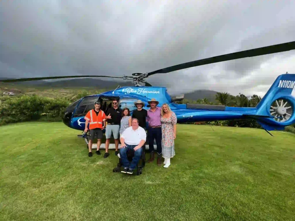 A group of seven people, including one person in a wheelchair, posing together in front of a bright blue 'Blue Hawaiian' helicopter on a grassy field with mountains and cloudy skies in the background.
