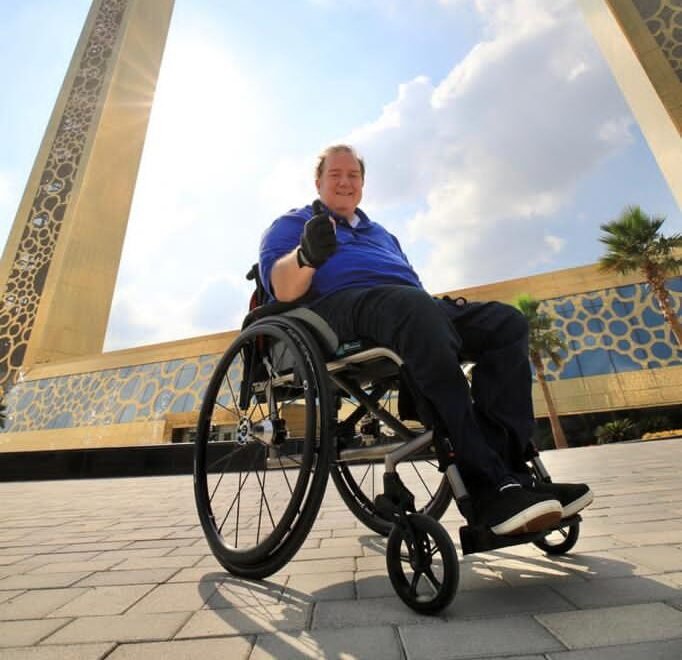 A man in a wheelchair wearing a blue shirt smiles and gives a thumbs-up in front of the Dubai Frame, a massive golden rectangular landmark, with the sun shining through partly cloudy skies.