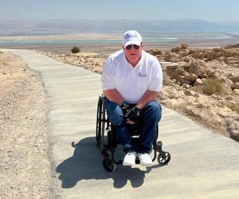 A man in a wheelchair wearing a white polo shirt and cap on a paved path at Masada, Israel, with desert landscape and the Dead Sea visible in the background.