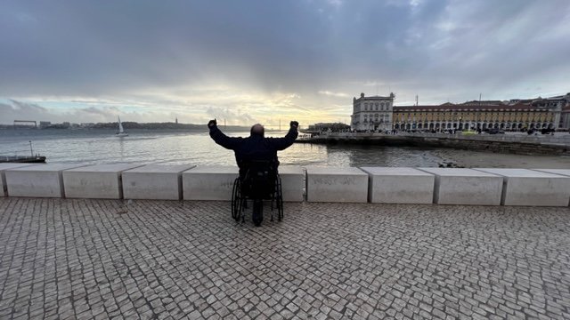 A person in a wheelchair facing the Tagus River in Lisbon at sunrise, raising their arms in triumph with the Praça do Comércio square and historic buildings in the background.