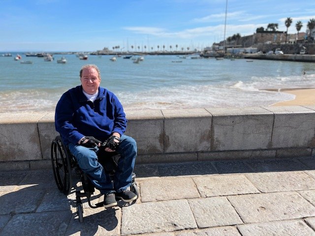 Man in wheelchair enjoying a sunny day by the seaside promenade in Cascais, Portugal, with boats on the water and palm trees in the background