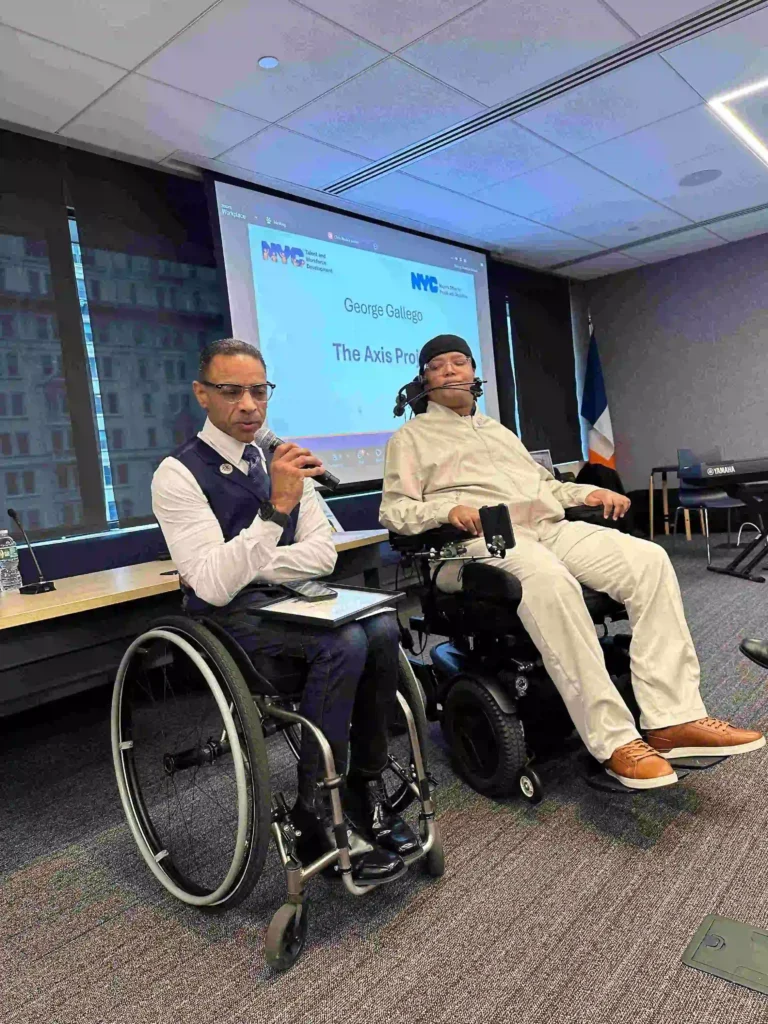 Two men in wheelchairs speaking at an event. The man on the left holds a microphone and tablet, while the man on the right sits in a motorized wheelchair with adaptive controls. Behind them, a screen displays the title 'George Gallego – The Axis Project' alongside NYC Talent and Development logos
