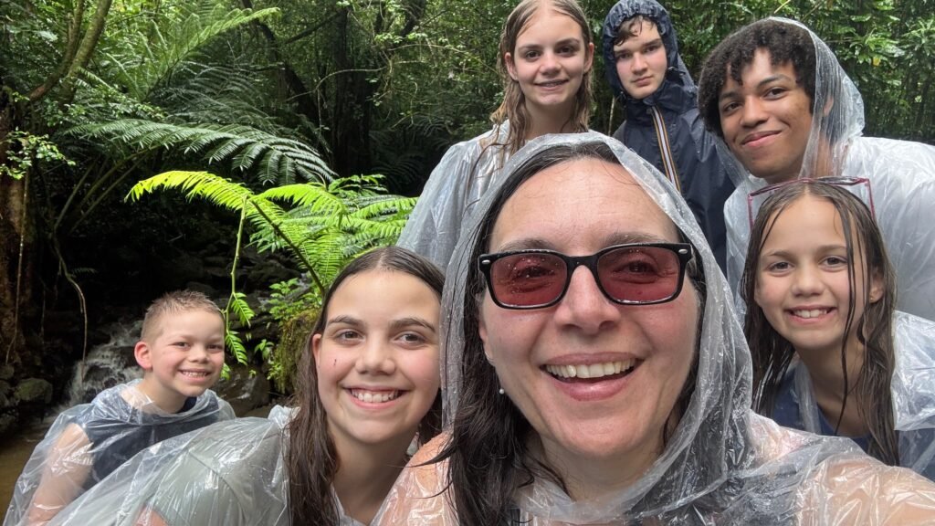 A group of people wearing clear rain ponchos pose in a lush green forest, with ferns and a stream in the background.