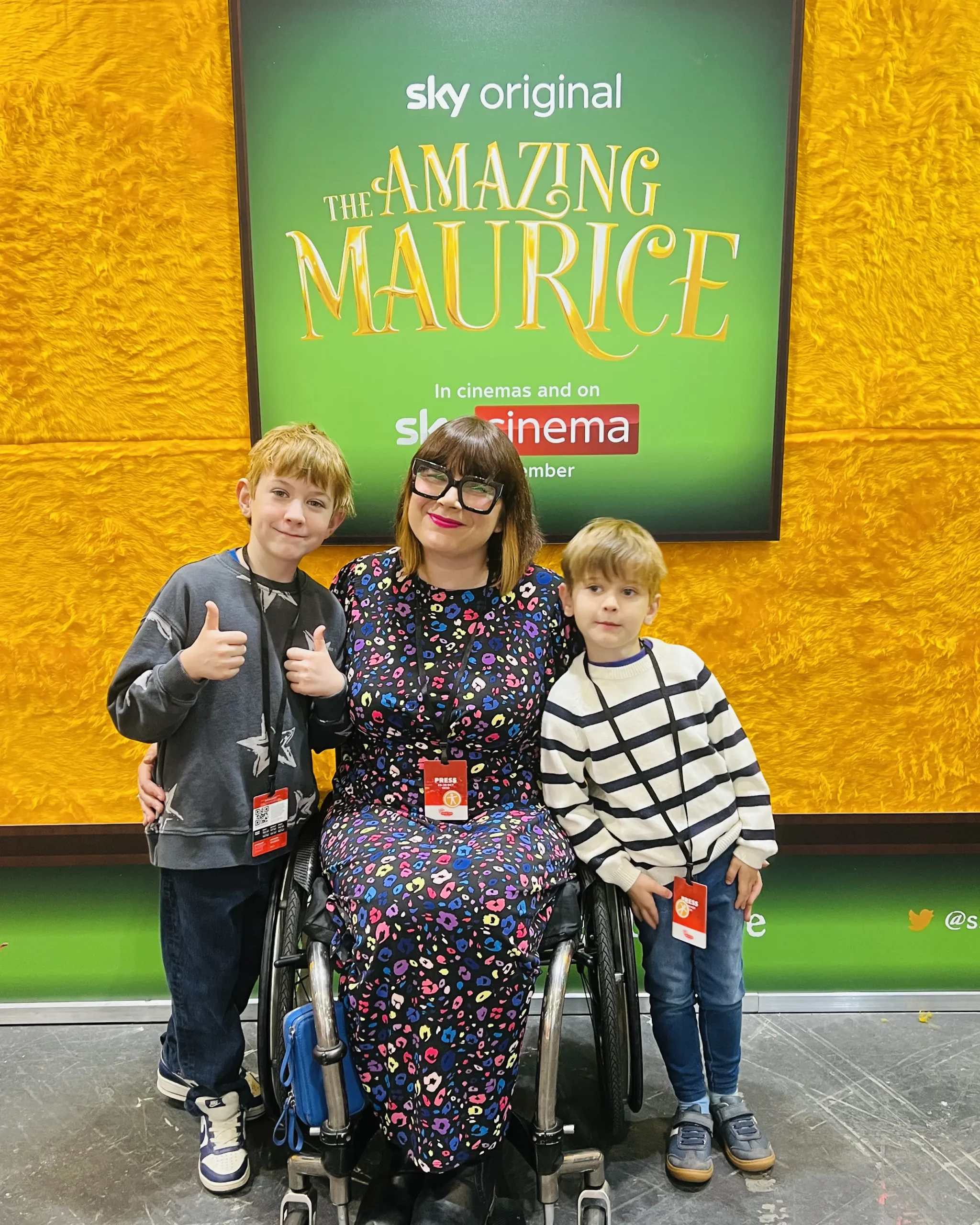 A woman in a wheelchair smiles at the camera, flanked by her two young sons. The three of them are in front of a movie poster for "The Amazing Maurice."