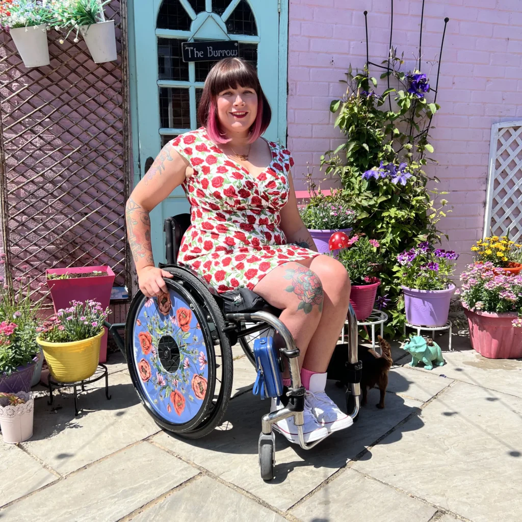 A woman with pink-tipped hair, in a wheelchair, smiles at the camera while sitting outside a pink house. She is wearing a white dress with a red rose pattern. A small black dog stands by her wheelchair.