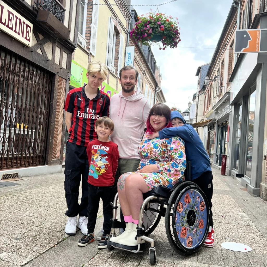 A family of five stands on a narrow cobblestone street lined with shops. The mother, in a wheelchair, has her arms around two of her sons. The father stands behind her with his arm on her shoulder, and the eldest son stands to the mother's left.