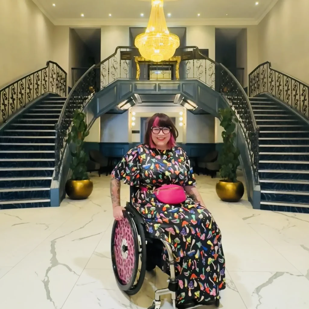A woman in a wheelchair is posing for a photo in a large, luxurious hotel lobby. She is wearing a long black dress with a colorful, whimsical pattern and a bright pink fanny pack. Behind her is a grand staircase and a large chandelier.
