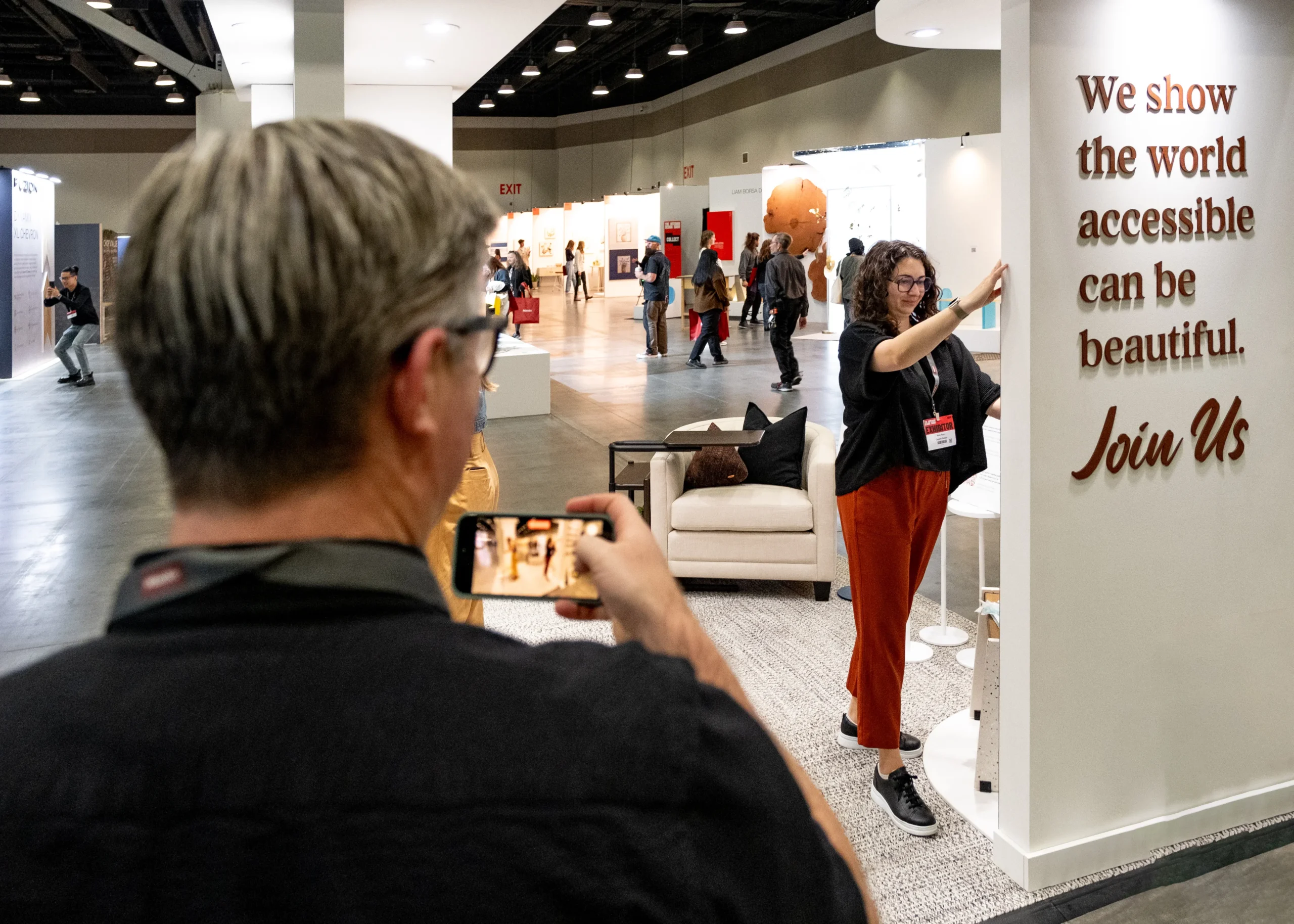 A man captures a photo of a woman posing next to a wall sign that reads, "We show the world accessible can be beautiful. Join Us."