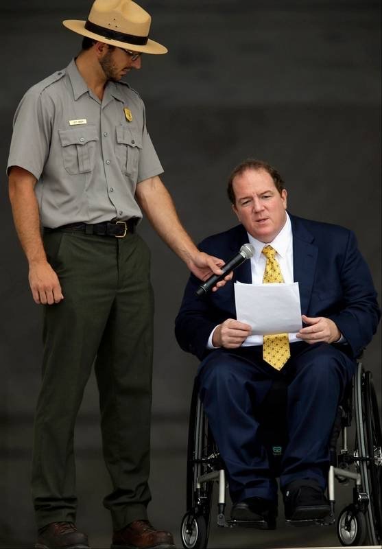 A man in a wheelchair wearing a suit and yellow tie speaks while holding papers, as a park ranger in uniform holds a microphone for him during an outdoor event.