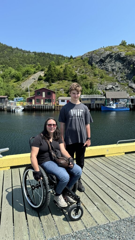 A woman in a wheelchair and a boy stand on a wooden dock with a scenic view of colorful houses and rocky hills in the background.