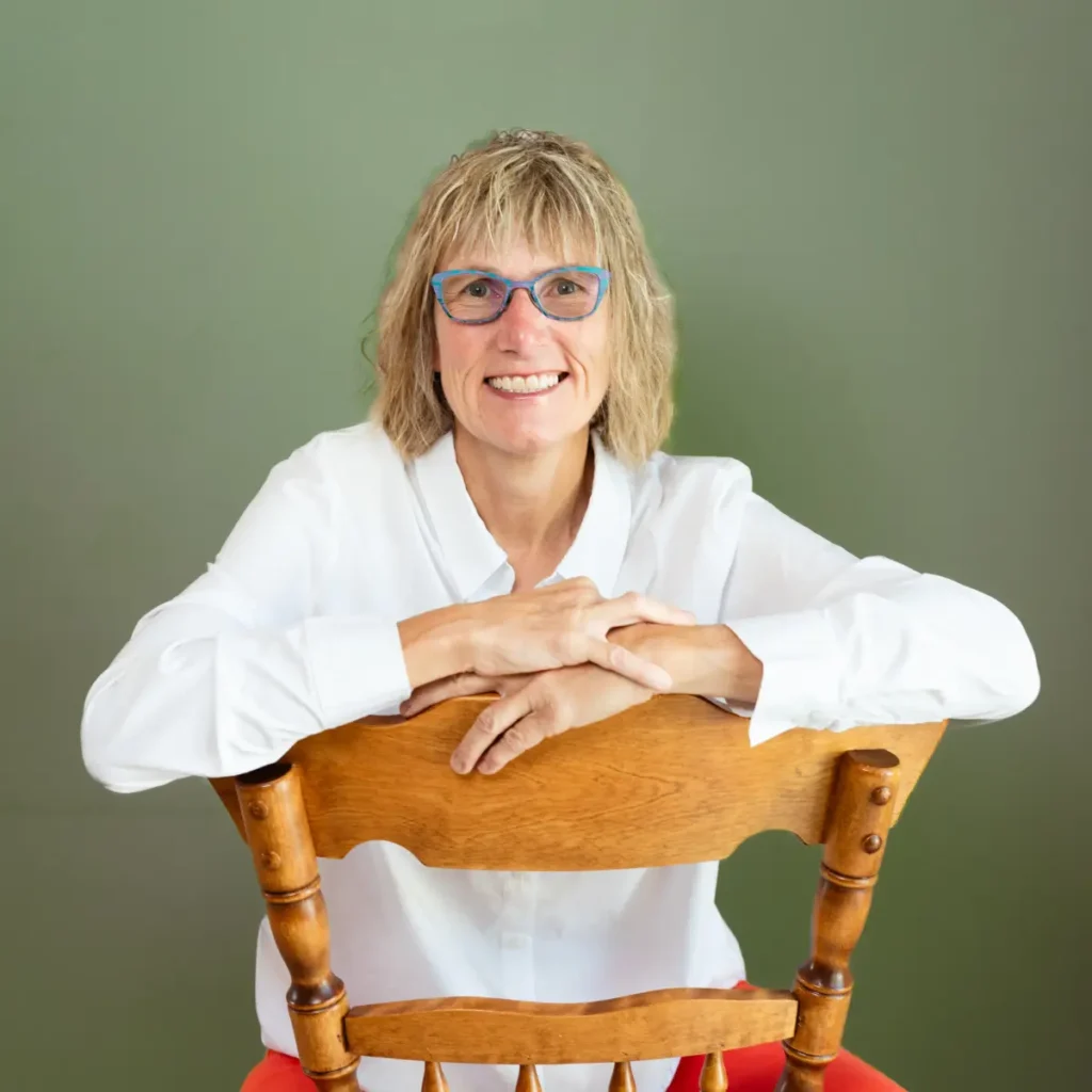 A headshot of Patti Bevilacqua smiling, leaning over the back of a wooden chair. She is wearing a white collared shirt and has her hands clasped on the chair back. She is wearing glasses with blue frames. The background is a solid green wall