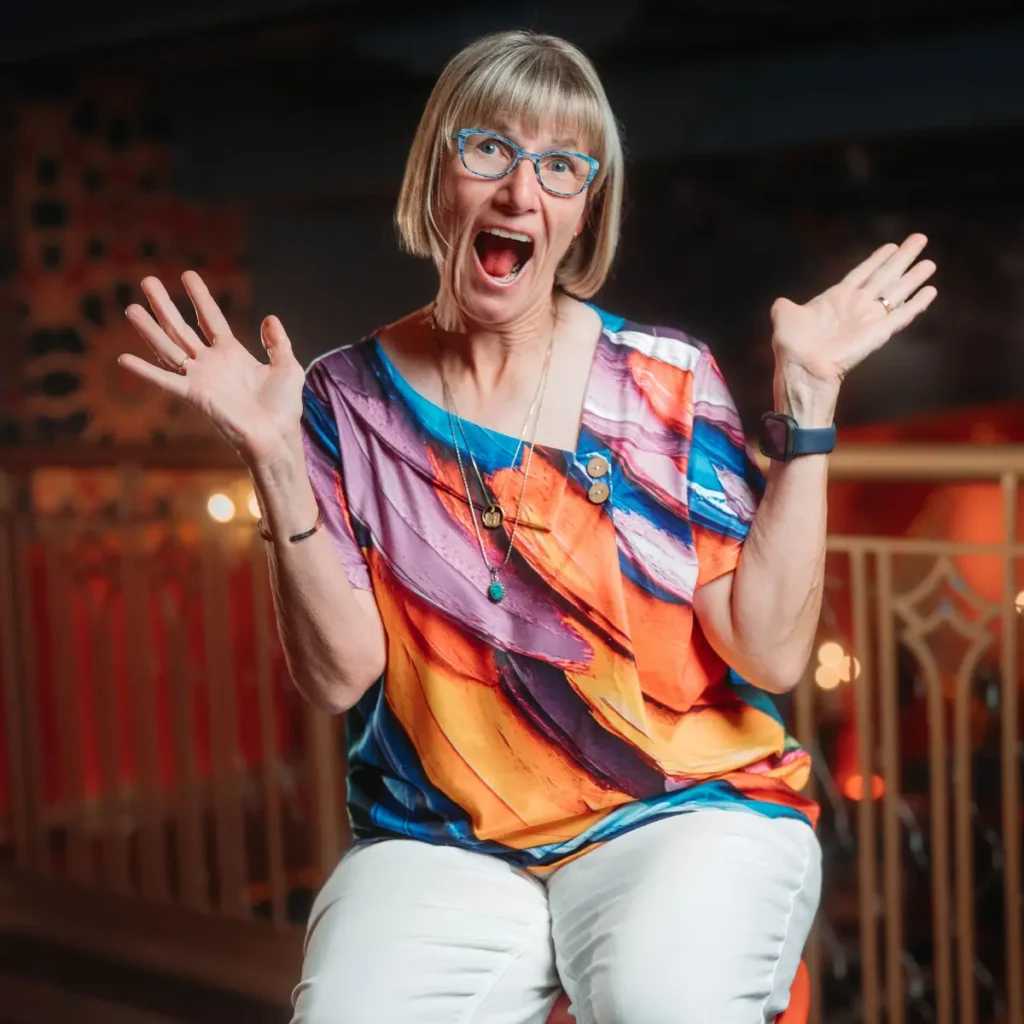 A portrait of Patti Bevilacqua in a state of excitement. She is seated, wearing a brightly colored abstract patterned blouse, white pants, and blue-framed glasses. Her mouth is open in a wide "O" shape, and her hands are raised in a gesture of surprise or amazement. The background is dimly lit with blurred warm lights.